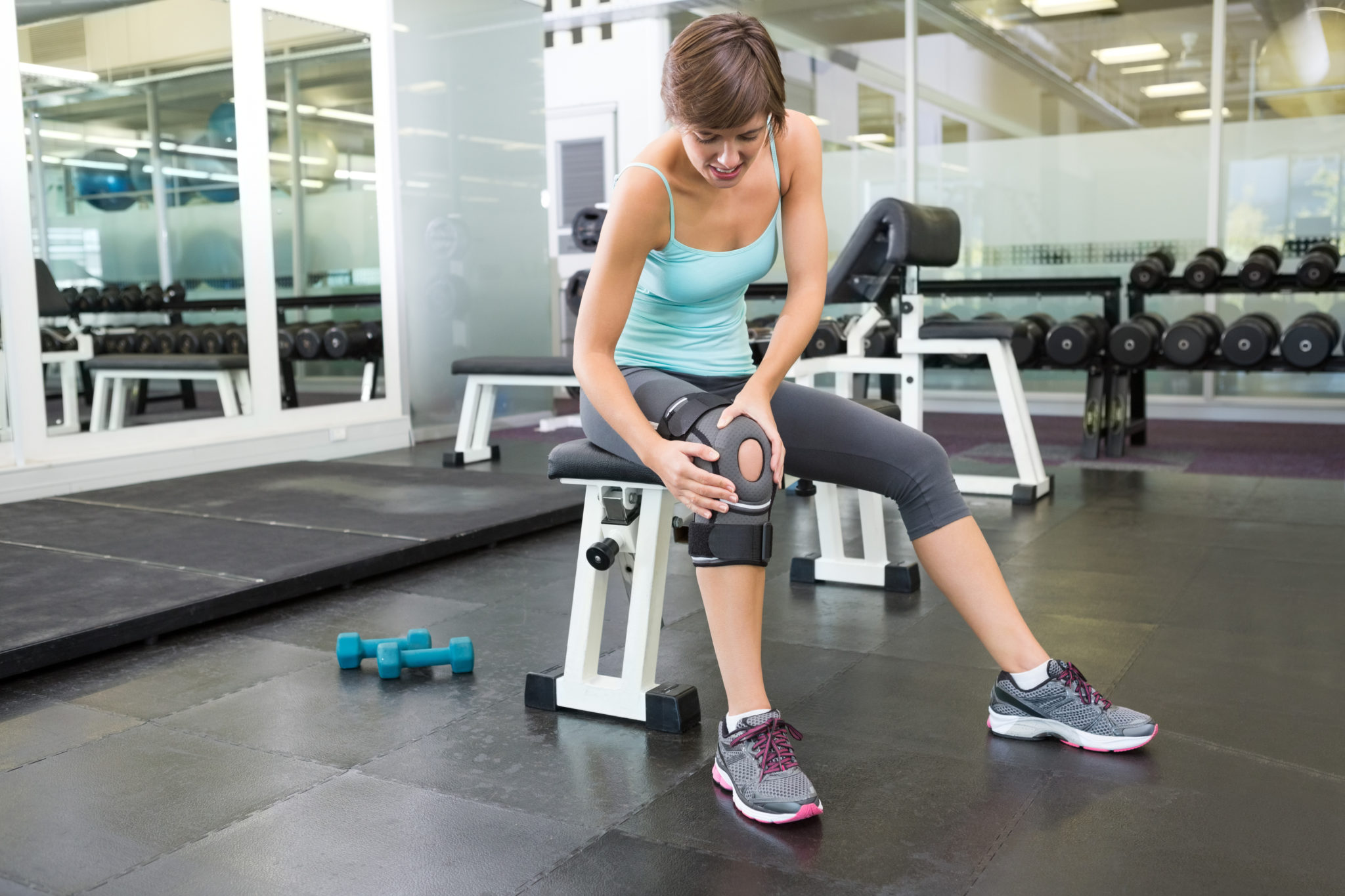 Fit brunette sitting on bench holding injured knee Fit brunette sitting on bench holding injured knee at the gym