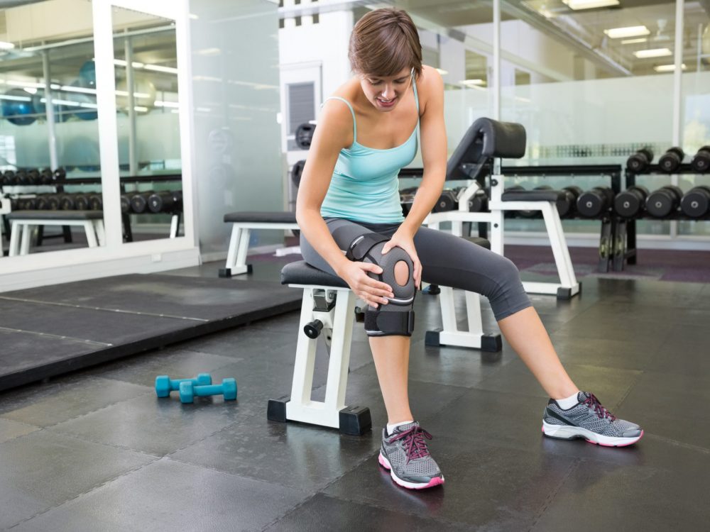Fit brunette sitting on bench holding injured knee Fit brunette sitting on bench holding injured knee at the gym